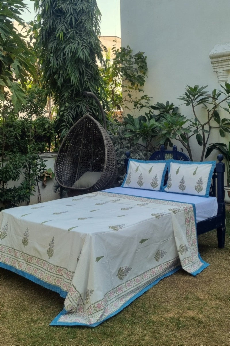 Hand block print cotton bedsheet with green floral motifs and blue border styled on wooden bed outdoors