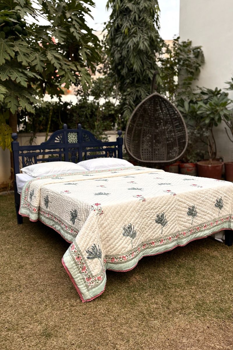 Hand block printed cotton quilt with green floral motifs and decorative border on a cream base, styled on a bed outdoors