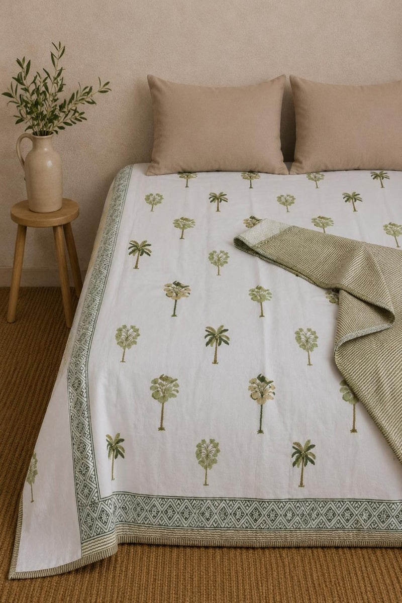 A bed dressed in a white cotton Dohar with a green palm tree pattern and a decorative border, styled with two beige pillows and a folded olive-green textured throw blanket. A small wooden stool with a vase of greenery sits beside the bed against a neutral beige wall.