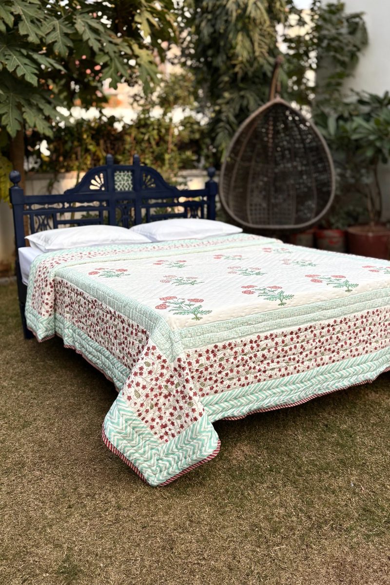 Hand block printed cotton quilt on wooden bed in garden setting, floral motifs with mint green border, artisanal Indian bedding