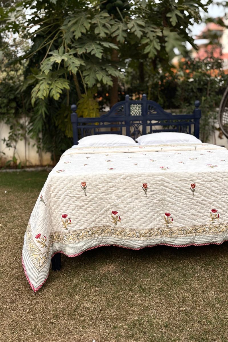 Hand block printed cotton quilt with red floral motifs and decorative border on a cream base, styled on a bed outdoors