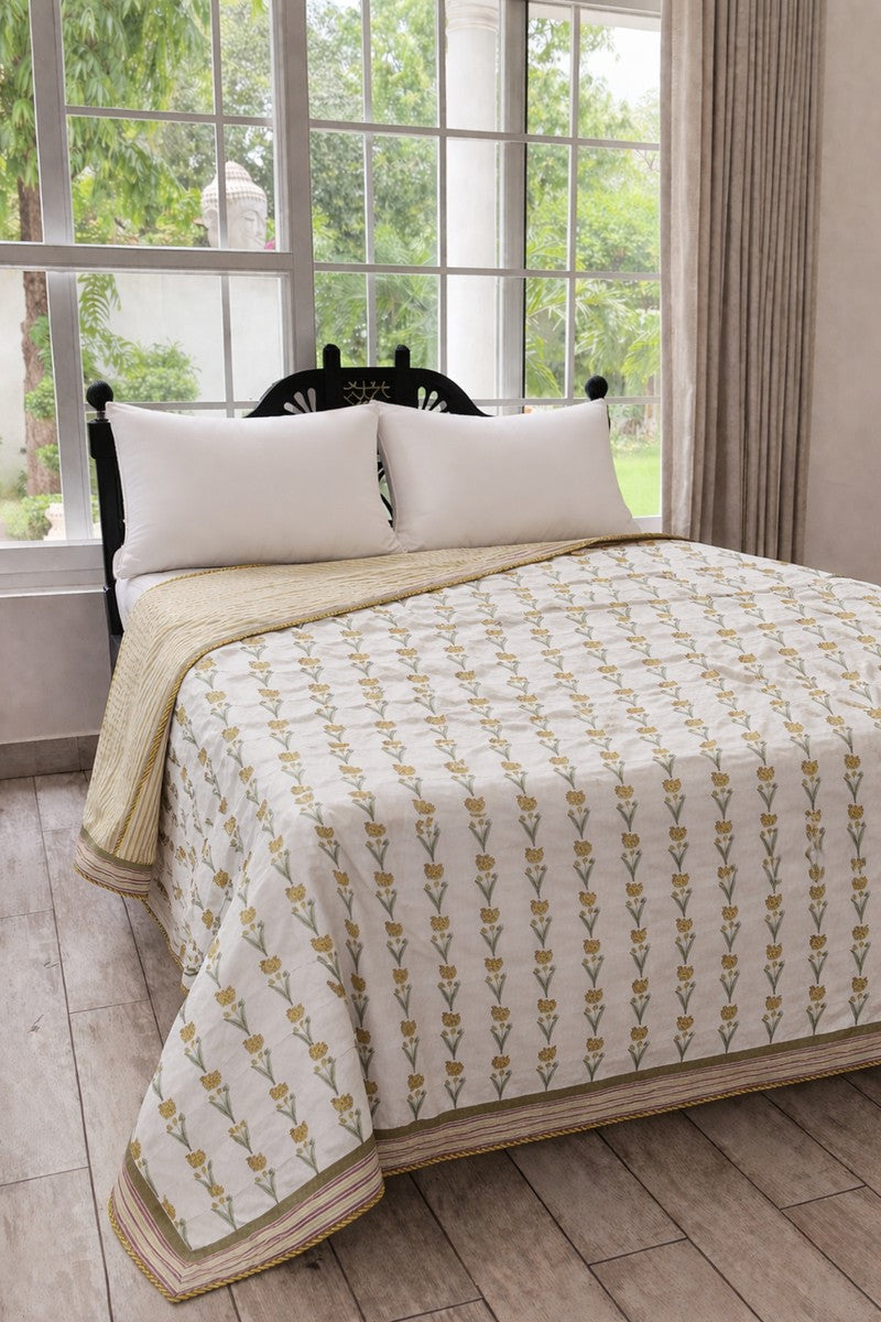 Bed with floral-patterned beige dohar and pillows in a room with large windows.