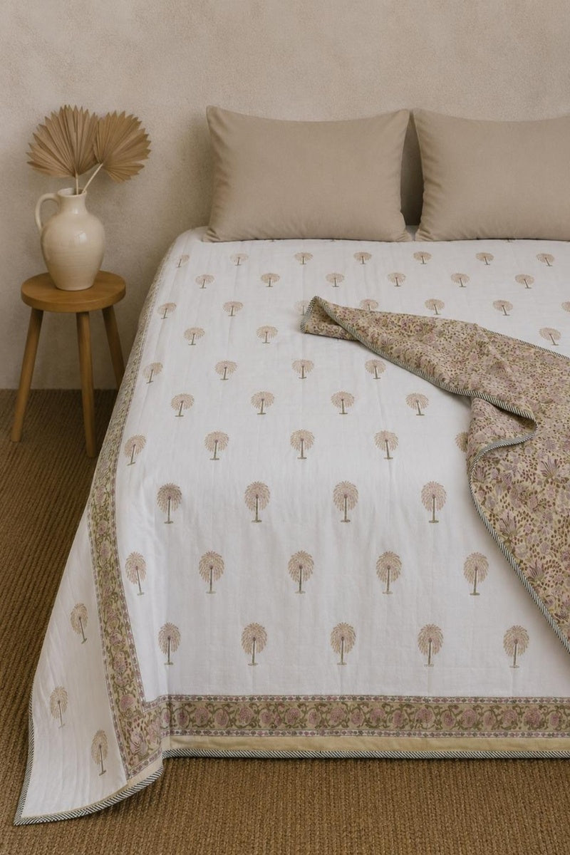 A white cotton Dohar with a beige tree pattern and floral border, styled on a bed with matching beige pillows and a folded patterned throw blanket. A small wooden side table with a vase is on the left.