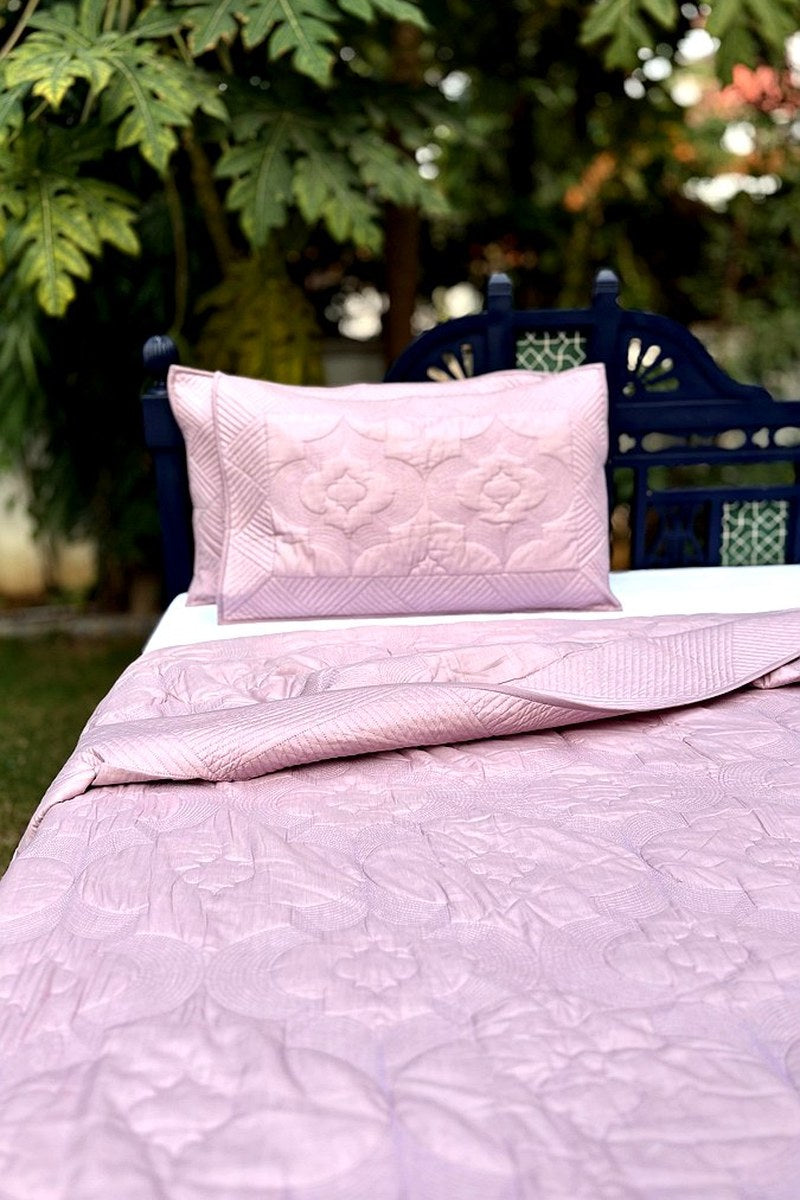 Luxury blush pink cotton quilt with intricate quilting, styled with matching pillows in a serene garden setting