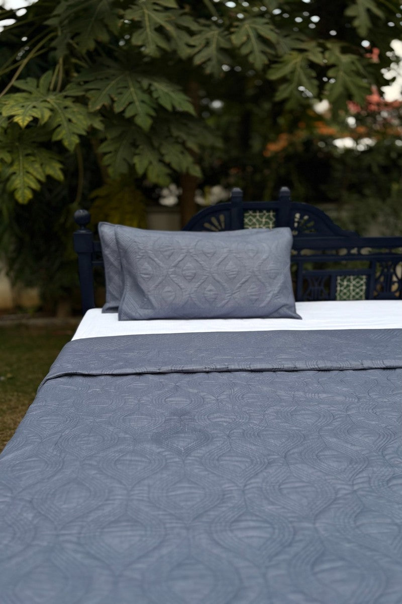Ash Mist grey quilted bedcover set styled on bed with navy headboard in garden setting