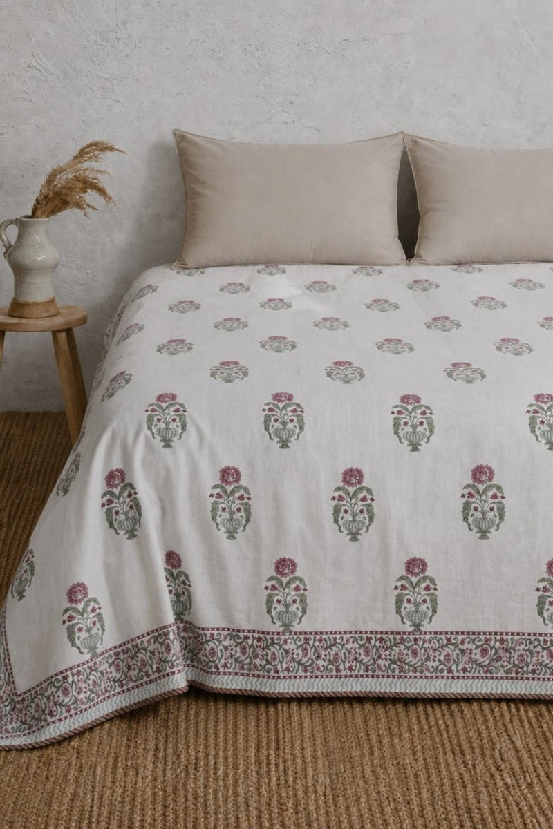 A white cotton dohar with a traditional block print pattern in muted green and dusty pink, styled on a bed with two beige pillows in a rustic bedroom setting.