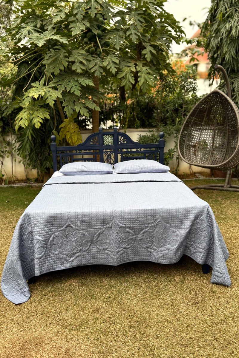Grey quilted cotton bedspread styled on wooden bed in a lush garden setting, featuring elegant geometric quilting and a soft matte finish