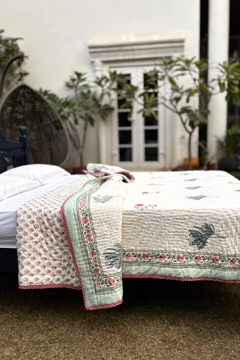 Traditional Indian hand block print quilt featuring floral motifs and soft green border, displayed on a wooden bed in a garden