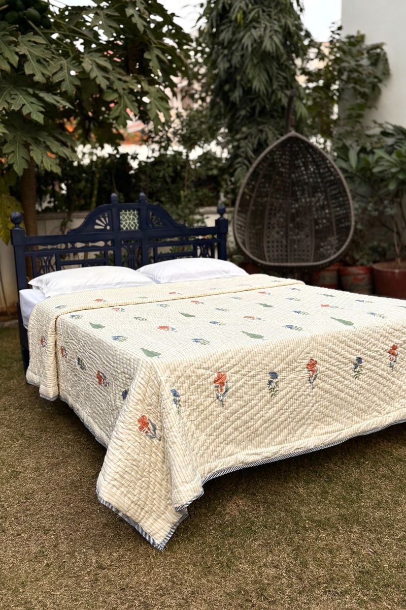 Hand block printed cotton quilt with multicolor floral motifs on a cream base, styled on a bed in a garden setting