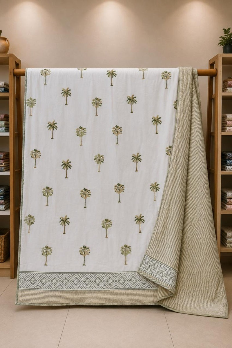 A white cotton Dohar featuring a repeating pattern of small green palm trees and a decorative border, draped over a wooden hanger in a home decor store.