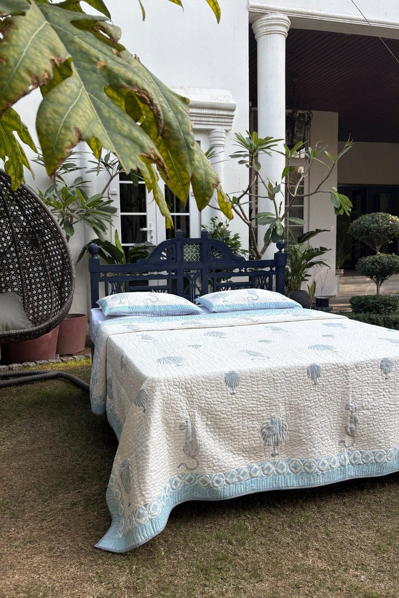 Blue and white hand block printed cotton quilted bedcover with pillows, palm tree and leopard motifs styled outdoors in garden setting