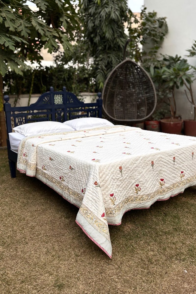 Traditional Indian hand block print quilt featuring small red floral motifs and ornate border, displayed on a wooden bed in a garden