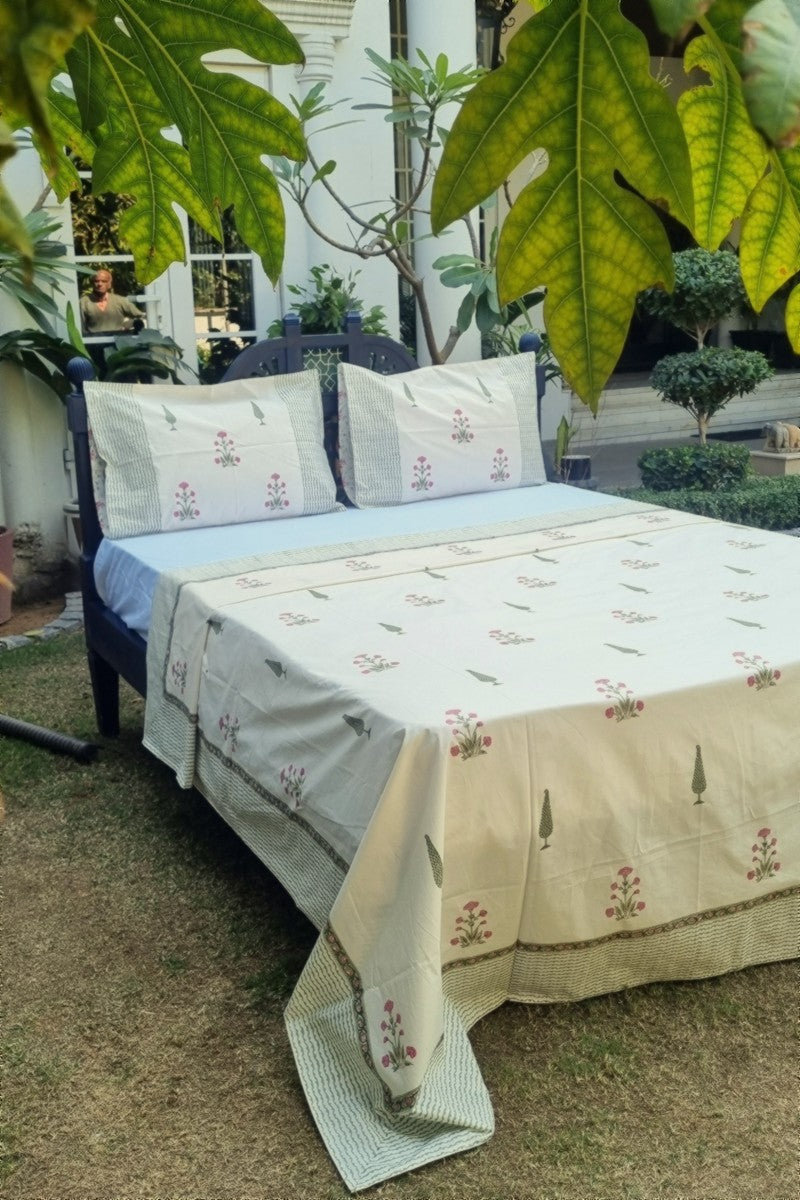 Hand block print cotton bedsheet set in ivory with pink floral motifs and green border styled on wooden bed outdoors