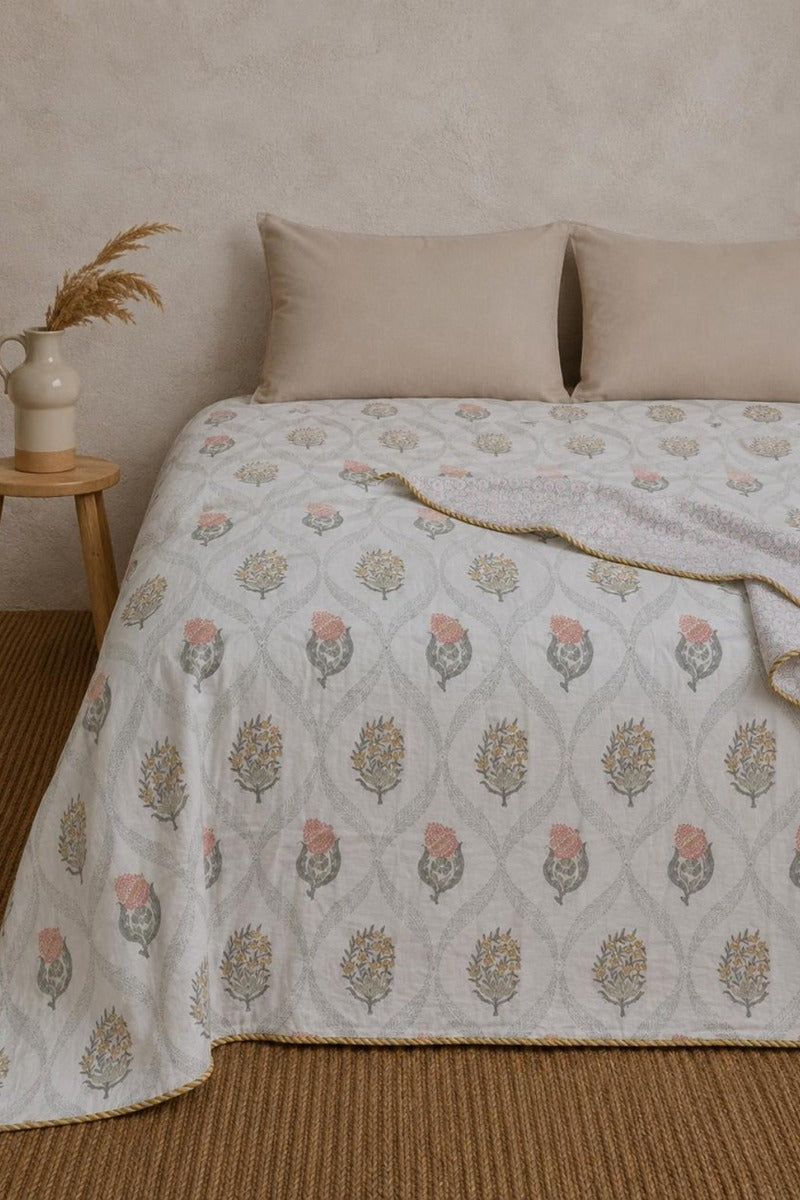 A light cotton dohar with a repeating floral and leaf pattern in muted pink, sage green, and gray, styled on a bed with two beige pillows in a rustic bedroom setting.
