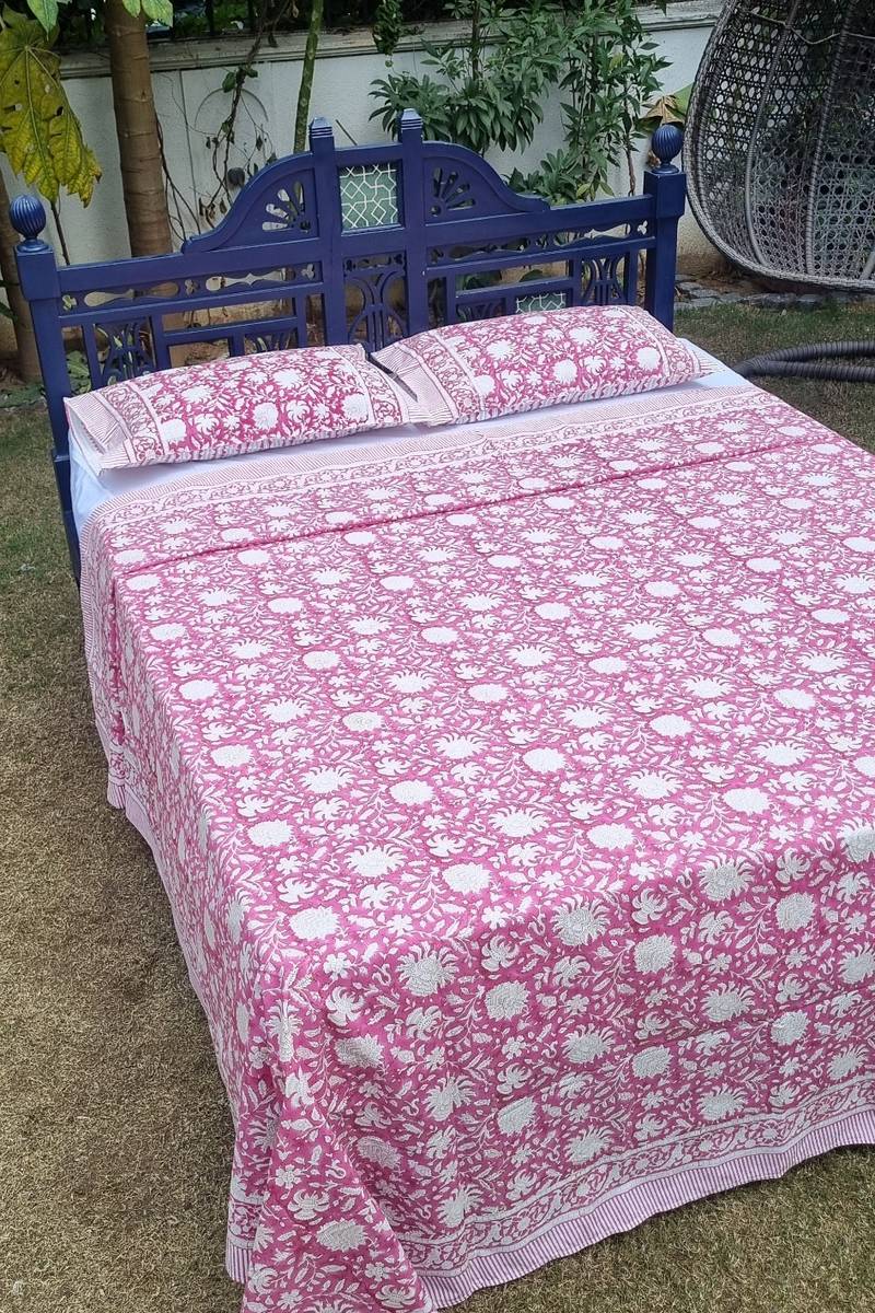Pink floral jaal print cotton bedsheet with coordinated pillow covers and border detailing styled on blue carved wooden bed in garden setting
