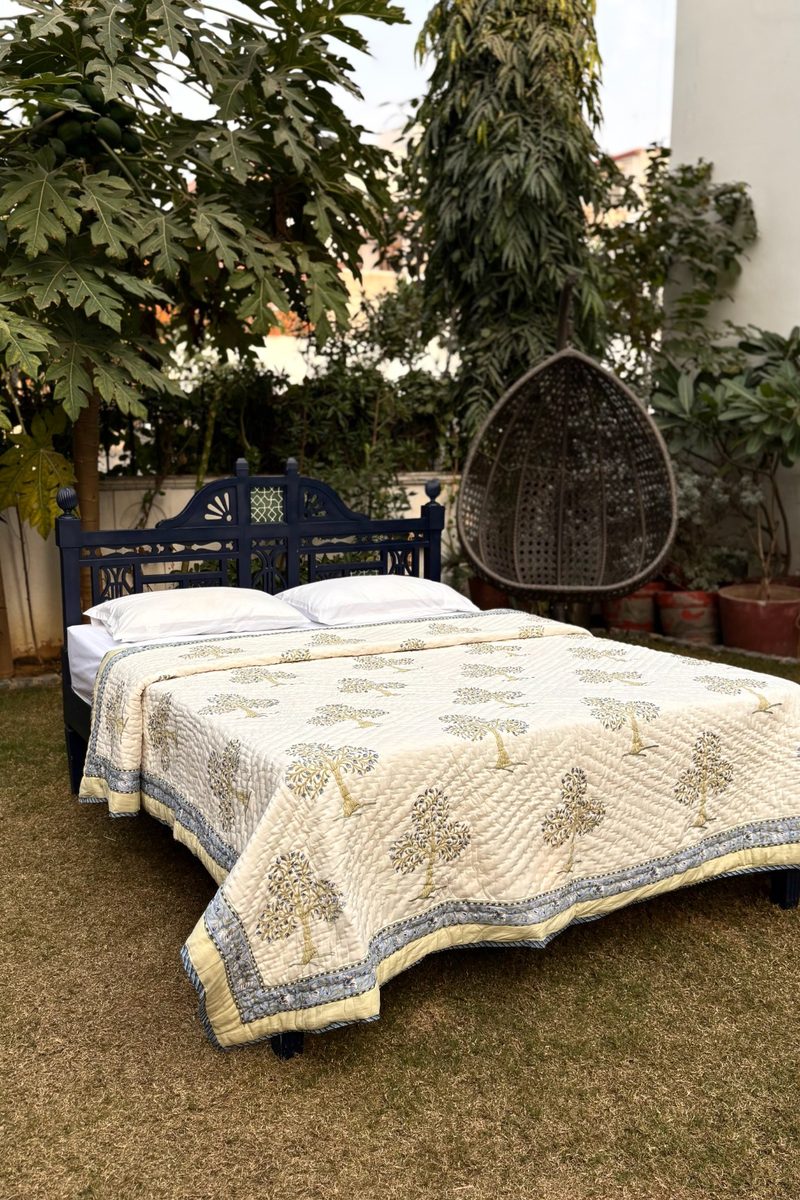 Cream and blue hand block printed cotton quilt styled on wooden bed, traditional tree design in outdoor setting